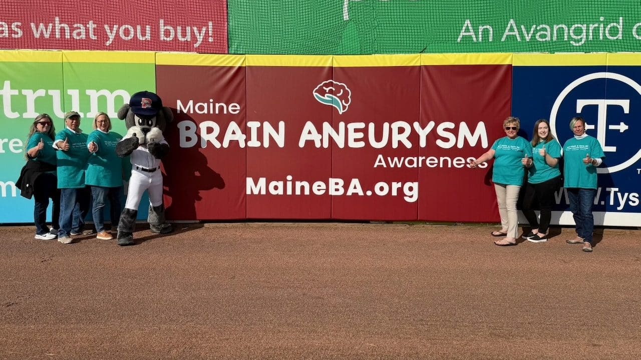 A group of people wearing teal shirts stands beside a mascot in a baseball stadium, promoting Maine Brain Aneurysm Awareness.